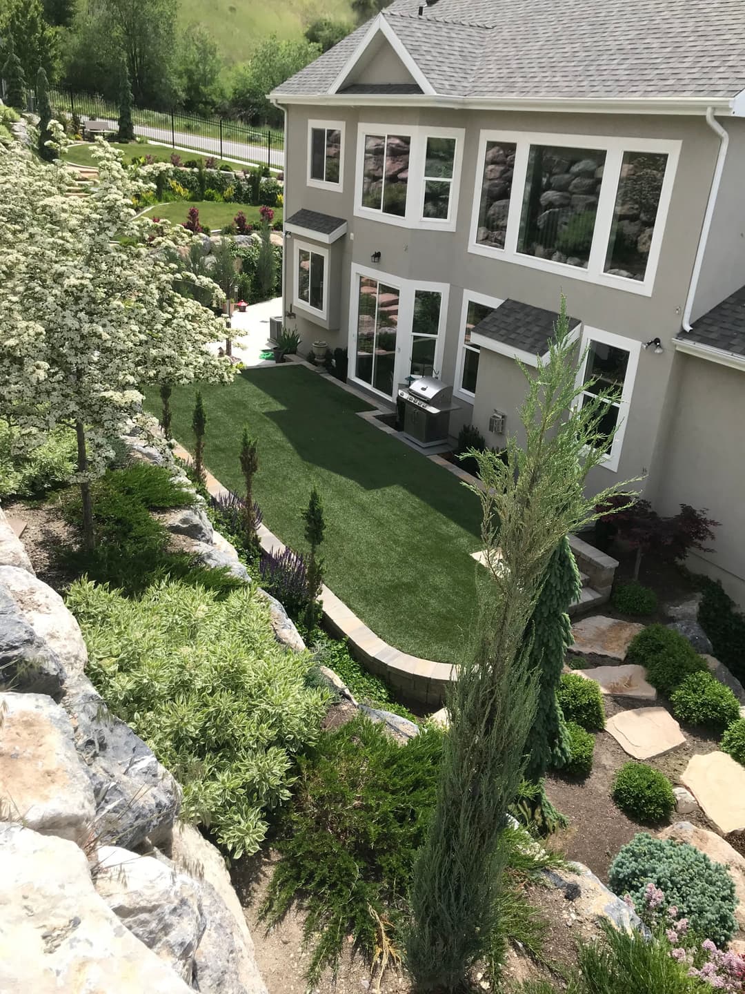 Lush backyard with artificial turf, landscaped plants, and a grill near a modern house.