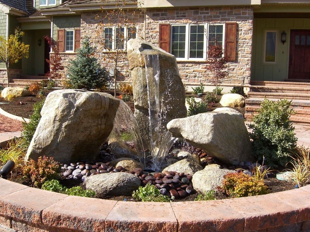 Natural stone fountain with water cascading over boulders in a landscaped garden setting.