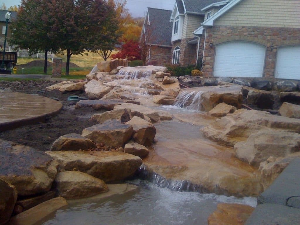 Stone waterfall feature in front of a residential home with autumn foliage.