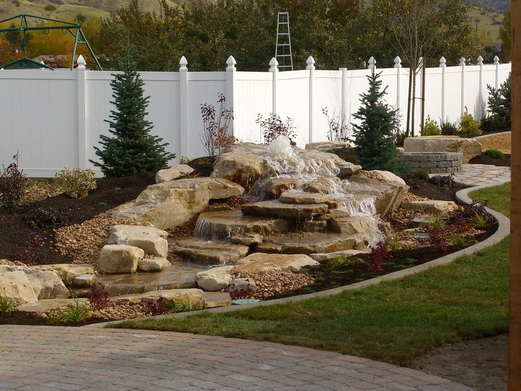 Backyard landscape featuring a stone waterfall, lush greenery, and white privacy fence.