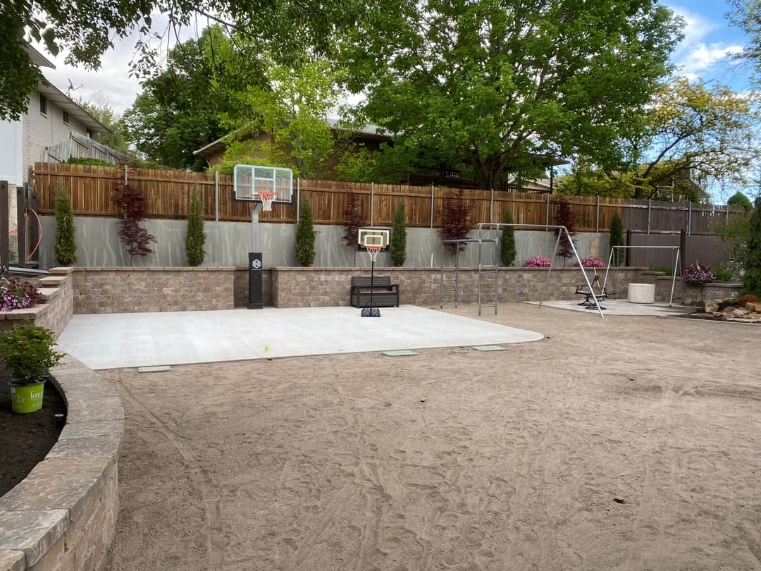 Backyard basketball court with a hoop, stone wall, and landscaping in a residential setting.