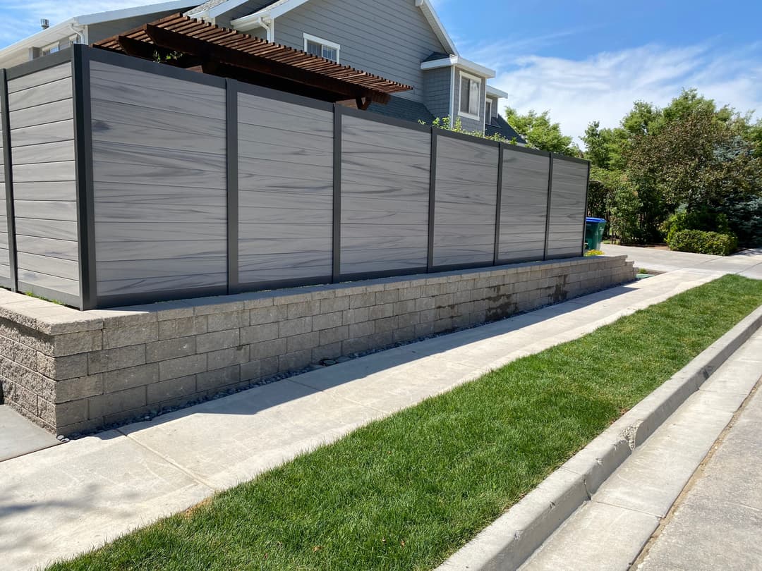 Modern privacy fence with wooden pergola, stone base, and manicured lawn beside a sidewalk.