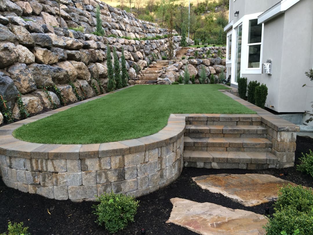 Landscaped backyard featuring artificial grass, stone stairs, and a rock wall.