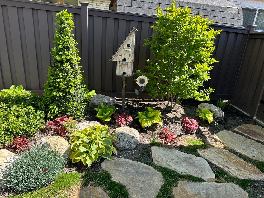Lush garden with birdhouse, hostas, and flowering plants beside stone path and fence.