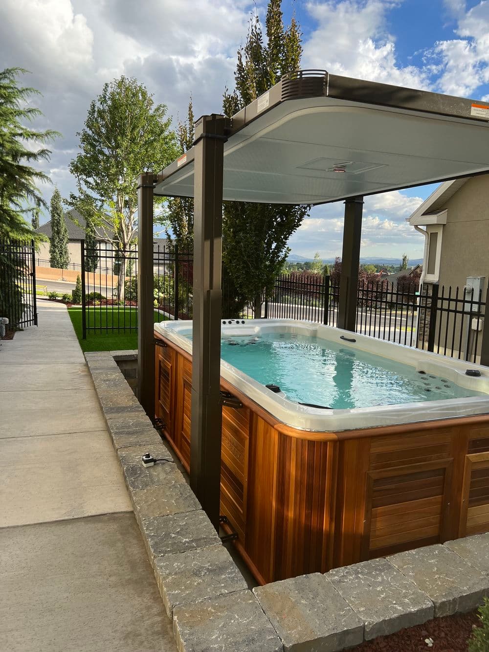 Outdoor hot tub with wooden paneling, covered by a metal gazebo in a landscaped yard.