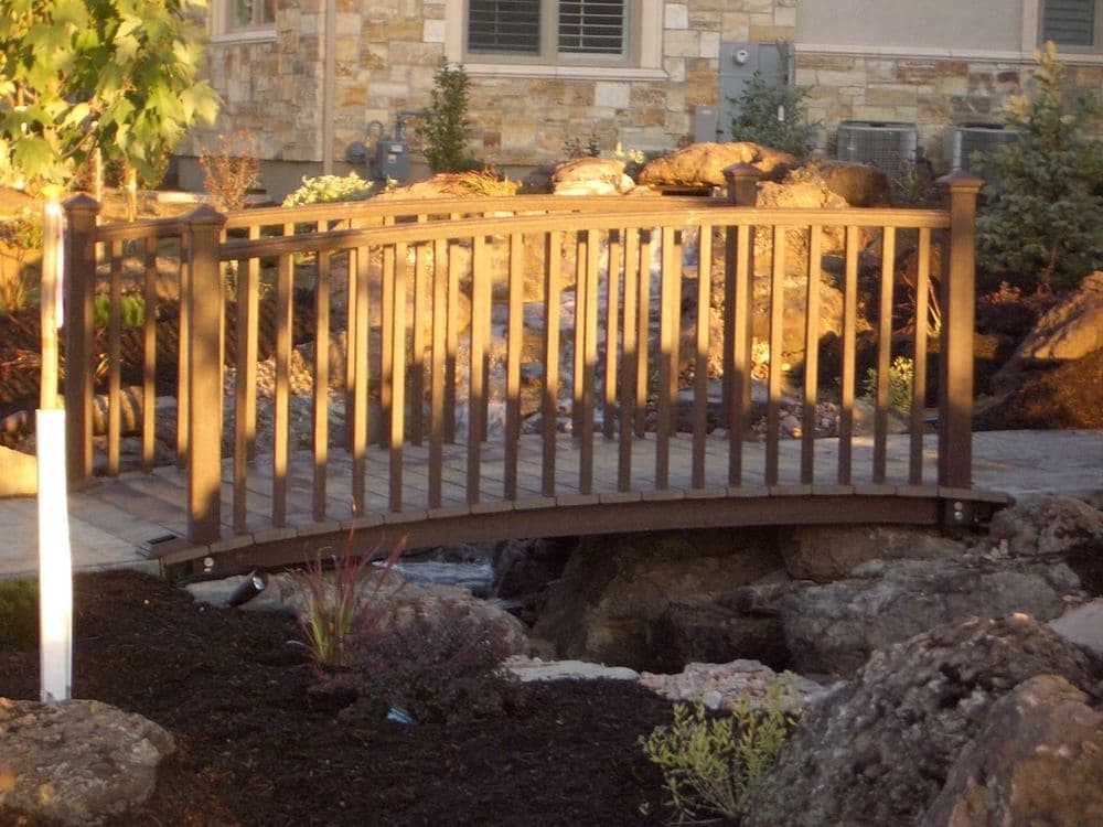 Wooden bridge arching over rocks in a landscaped garden with sunlight illuminating the scene.