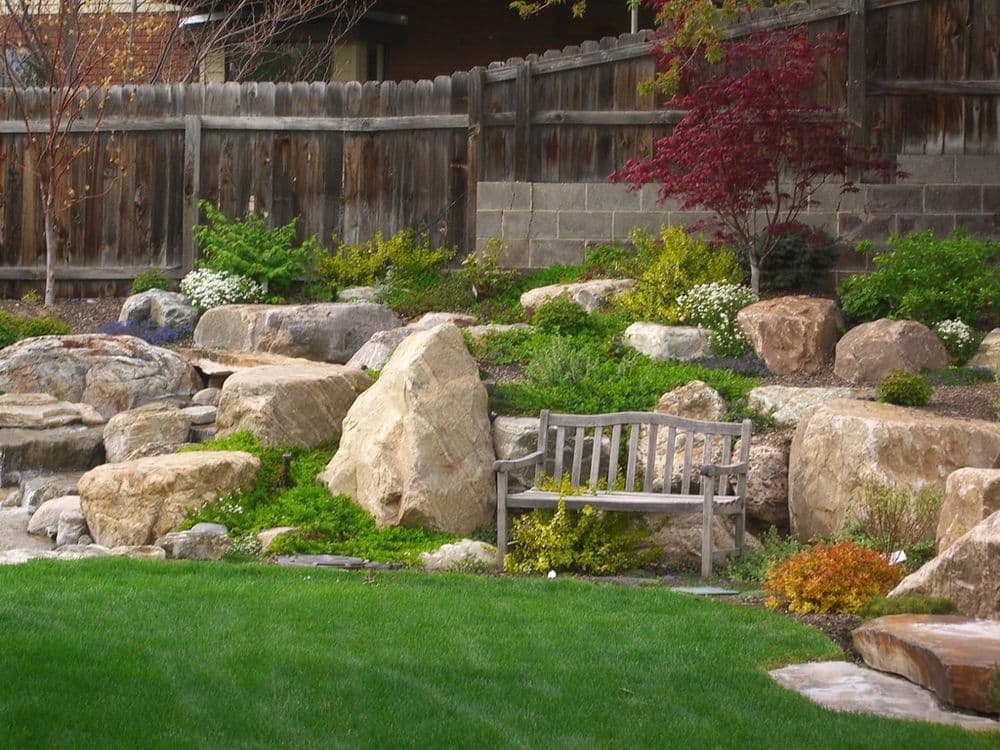 Serene garden with a wooden bench, rocks, and lush greenery in a landscaped yard.