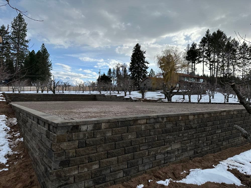 snow-covered landscape with a stone retaining wall and trees under a cloudy sky