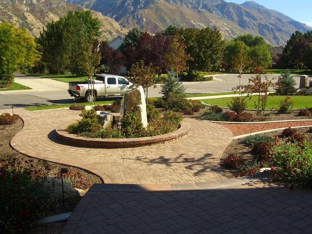 Landscaped pathway with stone features and flowers, surrounded by mountains and a parked truck.